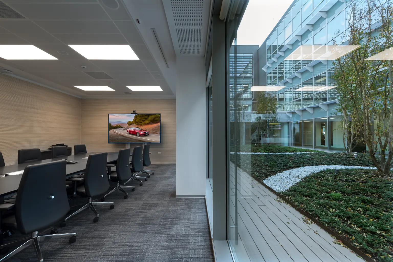 Una luminosa sala conferenze con un lungo tavolo e sedie da ufficio si affaccia su una grande finestra che dà su un cortile ben curato. Uno schermo sulla parete visualizza un'auto Mercedes-Benz.