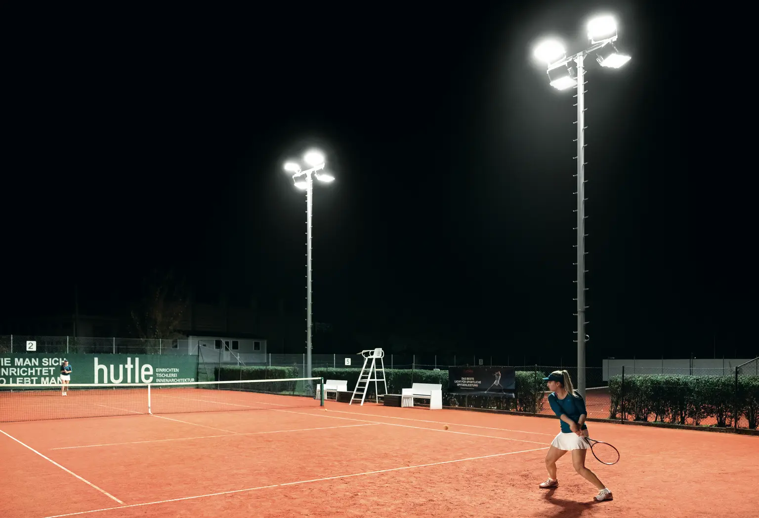 Ein Tennisspieler auf einem roten Sandplatz bei Nacht, der sich darauf vorbereitet, den Ball unter hellem Flutlicht zu schlagen. Der Platz ist von einem grünen Zaun, Sponsorenbannern und Bäumen im Hintergrund umgeben.