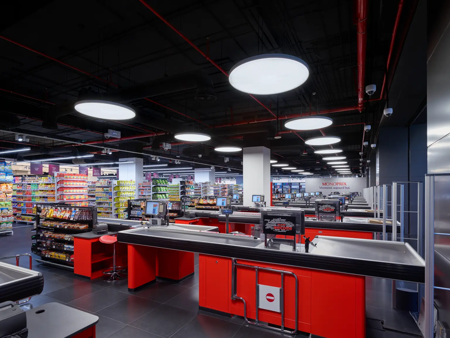 Un'ampia area casse in un supermercato con banchi rossi moderni, postazioni self-service e scaffali pieni di prodotti sullo sfondo.