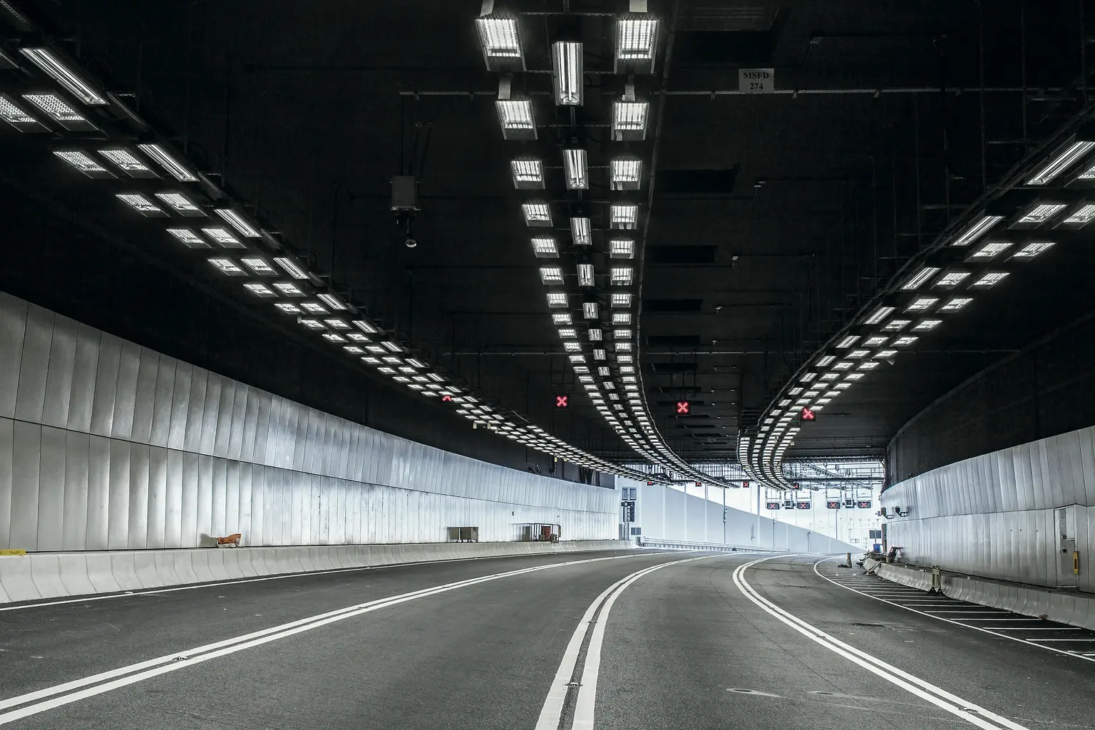 Un tunnel vuoto e ben illuminato con pareti in cemento liscio e luci brillanti sul soffitto che curva dolcemente verso sinistra. La strada ha chiare linee bianche di corsia e conduce verso l'uscita del tunnel.