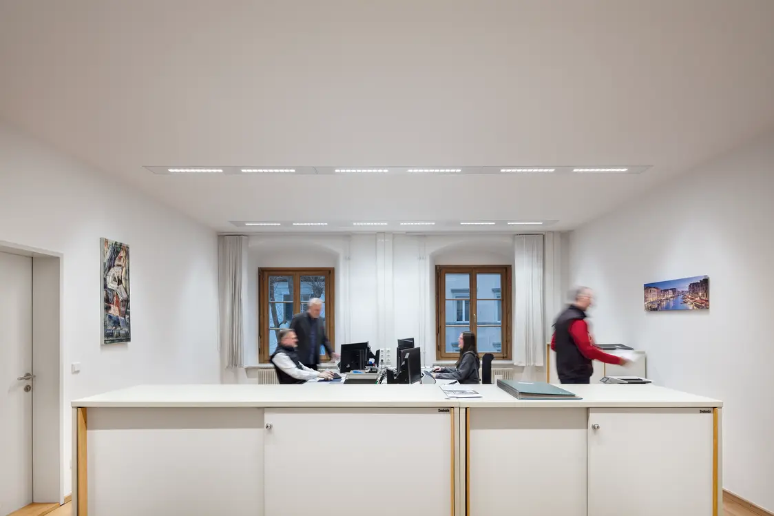 Helles Büro mit weißen Wänden, modernen Arbeitsplätzen und linearen LED-Leuchten in der Decke. Drei Personen arbeiten an Schreibtischen vor großen Fenstern, während an den Wänden Bilder hängen.