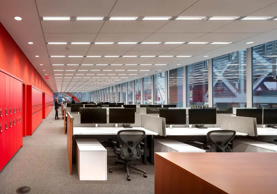 Ufficio open space con postazioni di lavoro moderne, dotate di scrivanie, monitor e sedie ergonomiche, oltre a una parete finestrata che lascia entrare molta luce naturale. Sullo sfondo si vedono due persone che conversano.