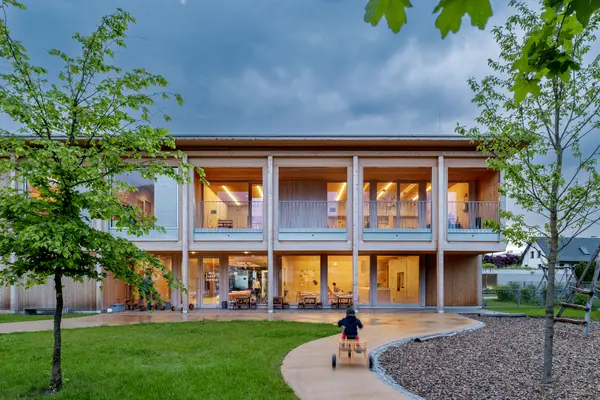  Casa bambini a due piani con facciata in legno, grandi finestre e illuminazione d'interni. L'architettura a misura di bambino unisce materiali naturali a una progettazione illuminotecnica invitante