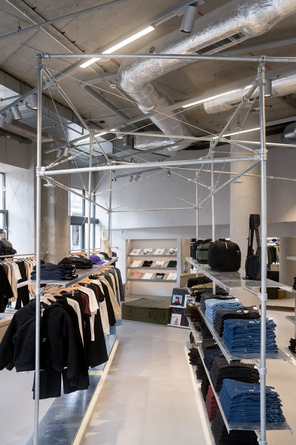 Carhartt WIP Store met industrieel ontwerp, zichtbare metalen structuren en ventilatiebuizen, modern verlichtingsconcept van Zumtobel. Op de voorgrond gevouwen jeans en kleding op kledingrekken, op de achtergrond schappen met accessoires en boeken.