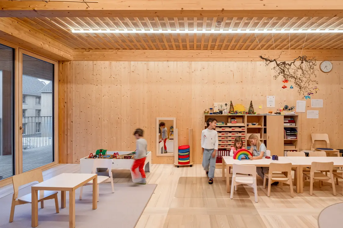 Area giochi nel centro per l'infanzia con rivestimenti in legno, arredi a misura di bambino e illuminazione uniforme a LED sul soffitto. La soluzione di illuminazione crea un'atmosfera tranquilla e sicura e supporta attività come giocare, fare lavoretti e apprendere