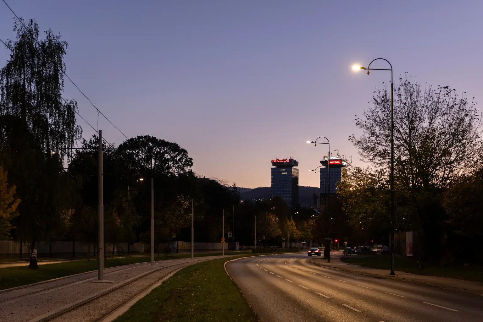Una strada periferica al crepuscolo, illuminata da lampioni curvi con alberi che fiancheggiano entrambi i lati. Due edifici alti con le cime illuminate di rosso sono visibili sullo sfondo sotto un cielo serale sfumato.