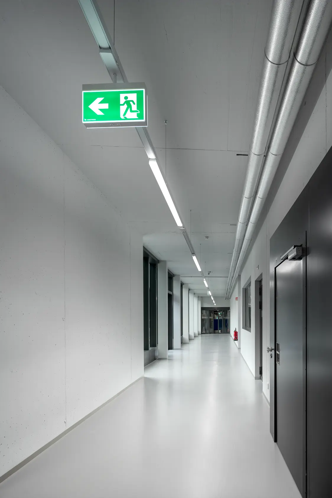Ein weisser Gang in der Industrieanlage. Ein Notausgangsschild hängt links oben an der Decke.