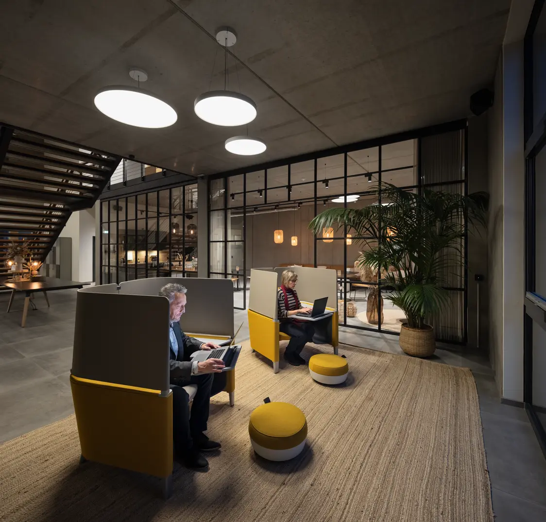 Twee personen werken in afgescheiden zithoeken onder ronde plafondlampen in een modern verlichte werkzone.