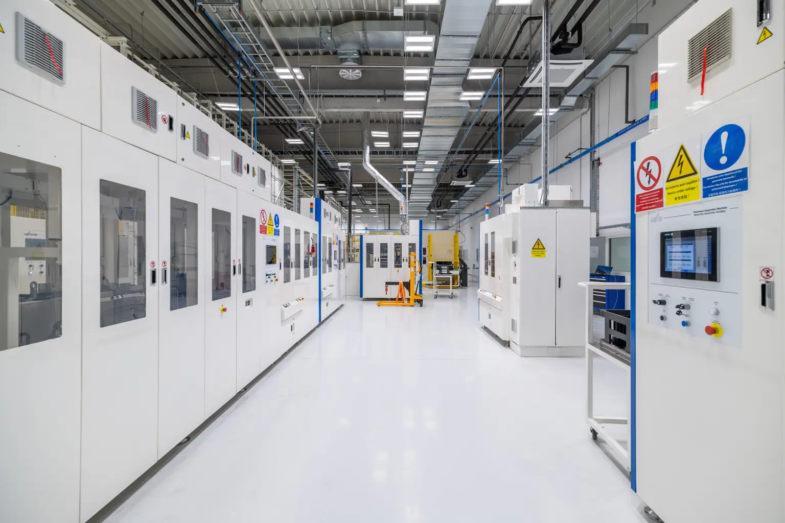An industrial production hall with white machines and blue columns, illuminated by bright LED lights on the ceiling. A yellow pallet truck is in the foreground, while the clear lighting highlights the technical details.
