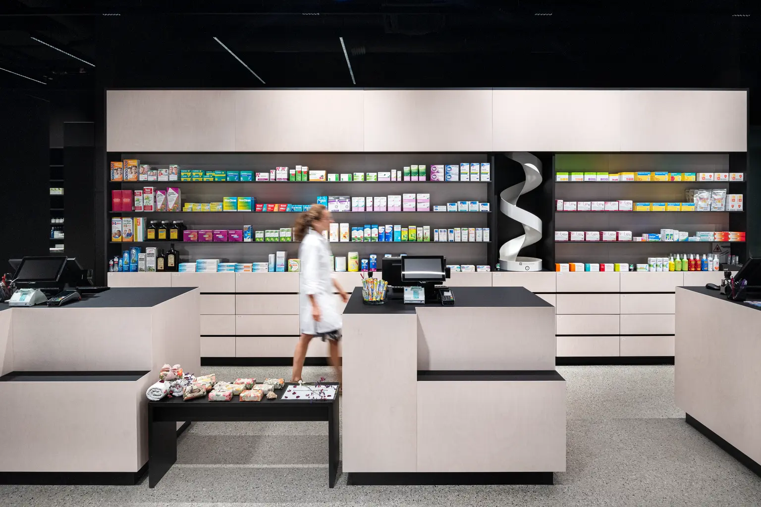 Una farmacia moderna con arredi luminosi e minimalisti e una struttura ordinata. Una dipendente si muove tra i banchi di vendita, mentre sullo sfondo sono visibili scaffali con medicinali e prodotti.