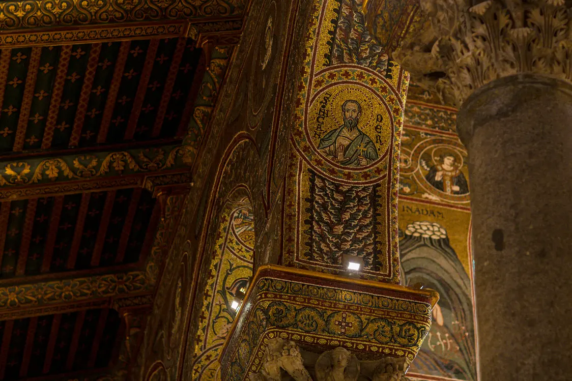 Detailaufnahme von reich verzierten Säulen und Bögen mit goldenen Mosaiken und Heiligenabbildungen in einer Kirche. Die Architektur zeigt kunstvolle Ornamente, religiöse Motive und eine aufwendig gestaltete Holzdecke.