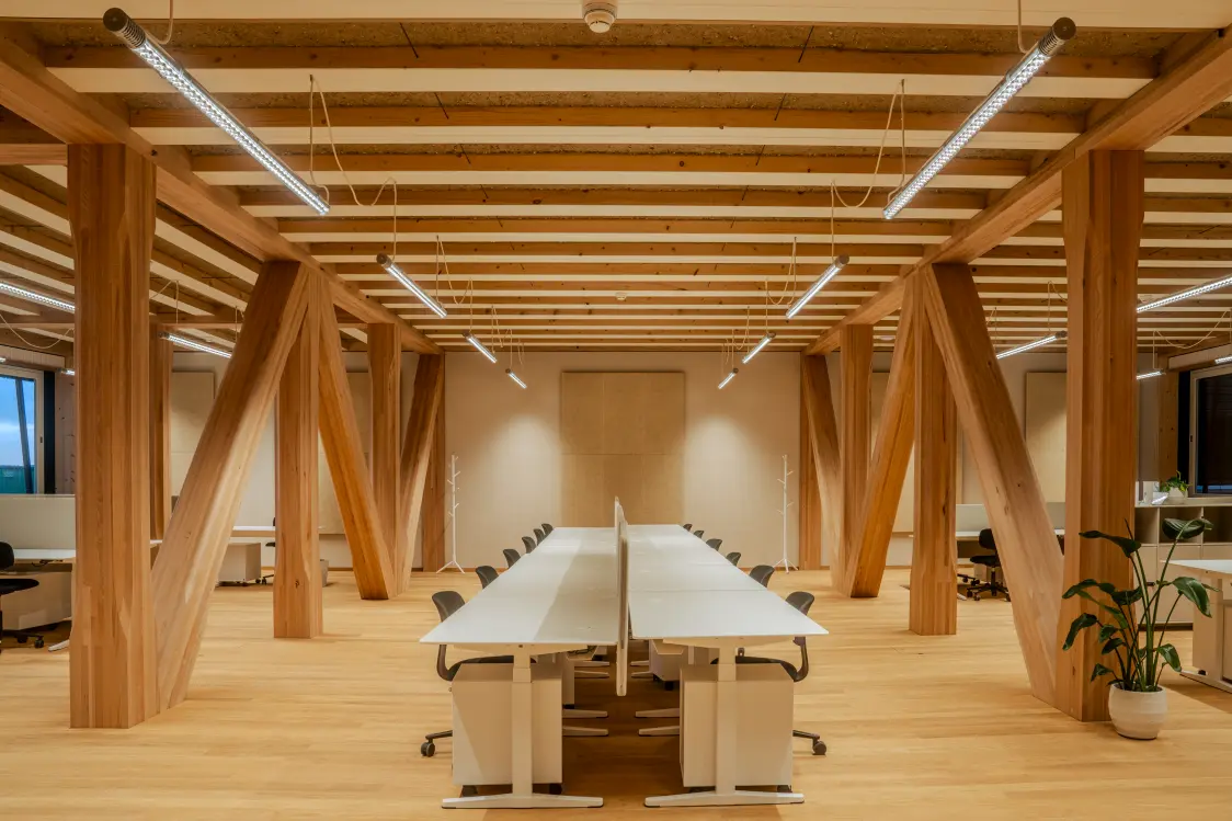 Vista interna di una moderna sala riunioni con schermo, tavolo e illuminazione minimalista, design sostenibile con tubature a vista e soffitto in legno.