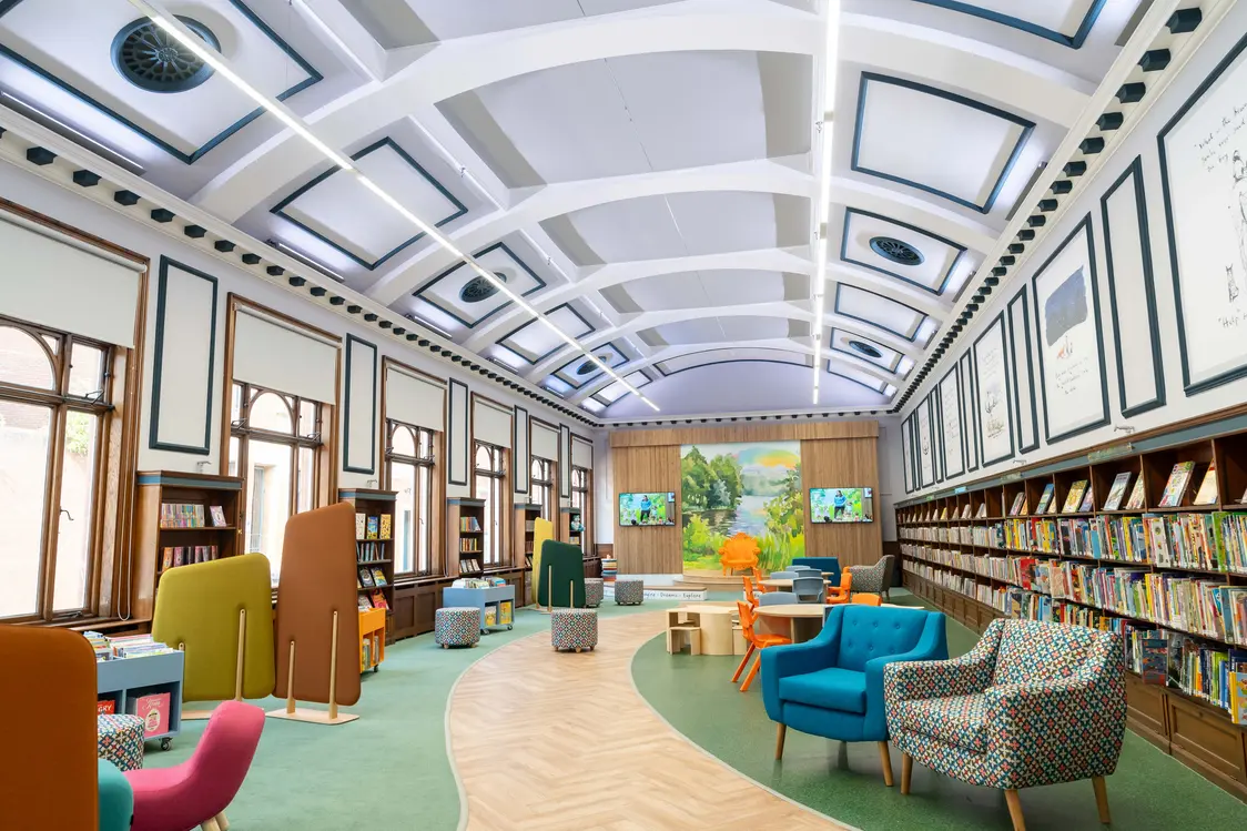 Ein heller Bibliotheksraum mit einer weißen Gewölbedecke, beleuchtet durch lange LED-Lichtleisten. Die Bücherregale und farbenfrohen Sitzmöbel schaffen eine einladende Atmosphäre, während ein Bildschirmbereich im Hintergrund zu sehen ist.