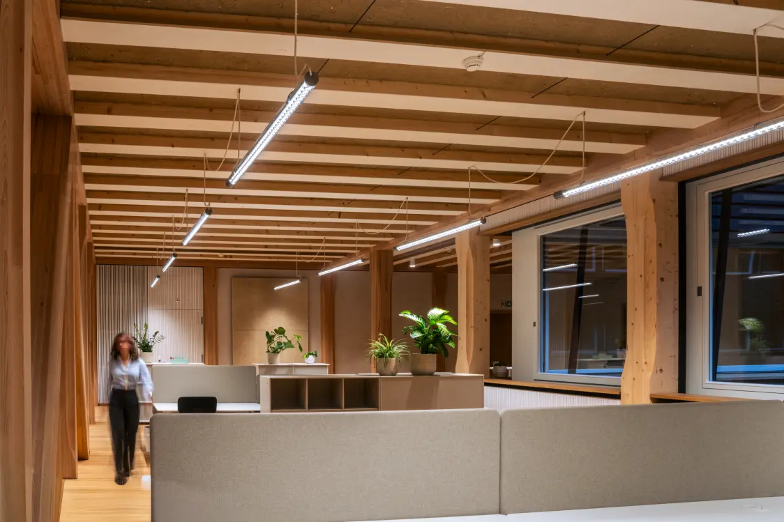 Vista interna di una moderna sala riunioni con schermo, tavolo e illuminazione minimalista, design sostenibile con tubature a vista e soffitto in legno.