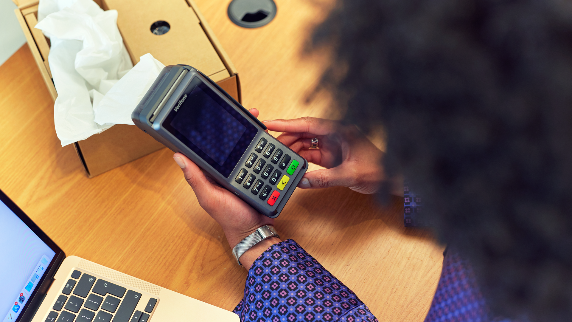 Pessoa fazendo um pagamento usando um terminal de cartão portátil ao lado de caixas de papelão e um laptop.