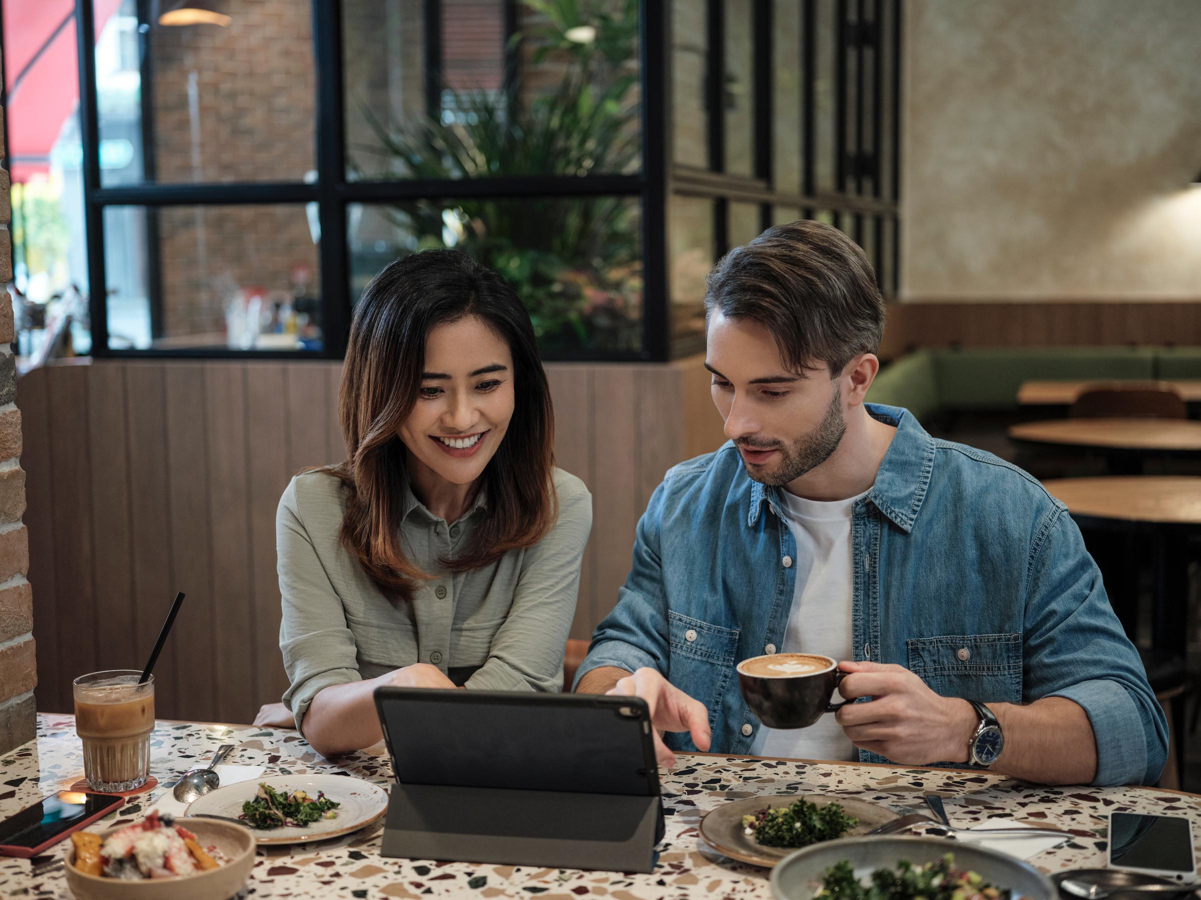 A man and woman sitting in a cafe, having coffee and lunch while looking at an ipad 