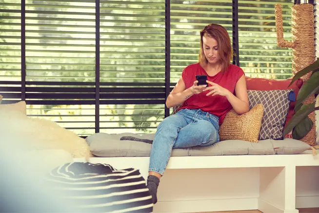 Mujer sentada en un sofá utilizando un smartphone con una planta y persianas en el fondo.
