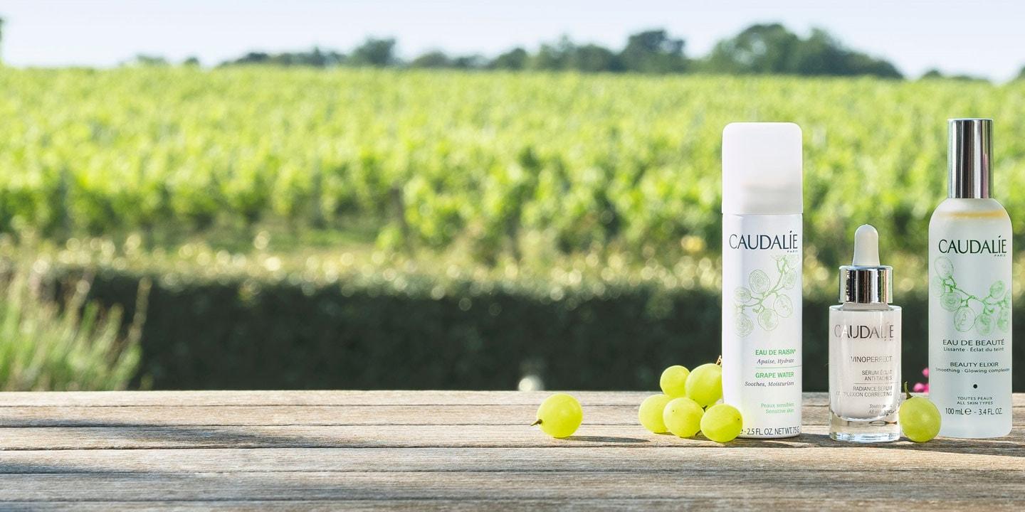 Produits de soins Caudalie et raisins sur surface en bois avec vignoble en arrière-plan.