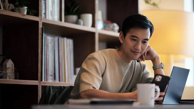Hombre sonriendo sentado con una laptop y sosteniendo una taza de café en una habitación acogedora con una estantería de libros.