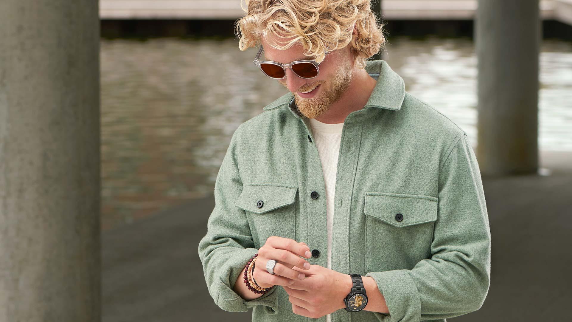 Leende man med lockigt hår som bär solglasögon, en grön jacka och en klocka.