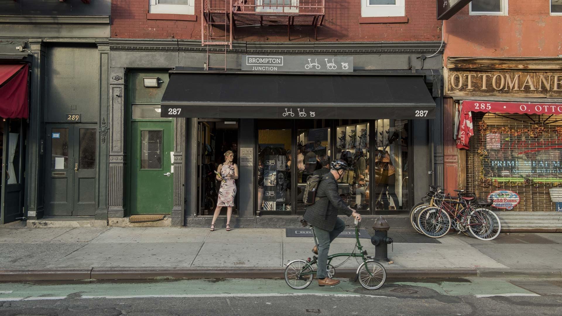 A cyclist passing by a Brompton bike shop with a woman standing at the door.
