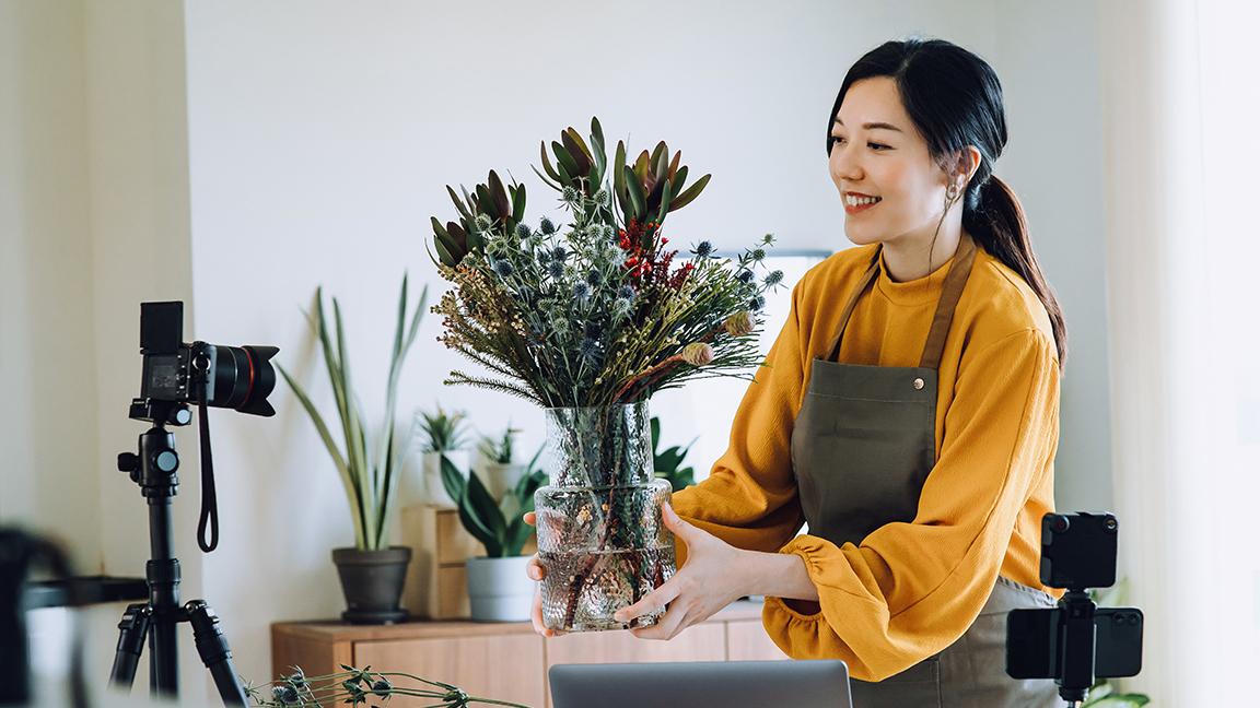 Smiling woman in yellow blouse and apron arranging flowers in a vase, with cameras on tripods in foreground.