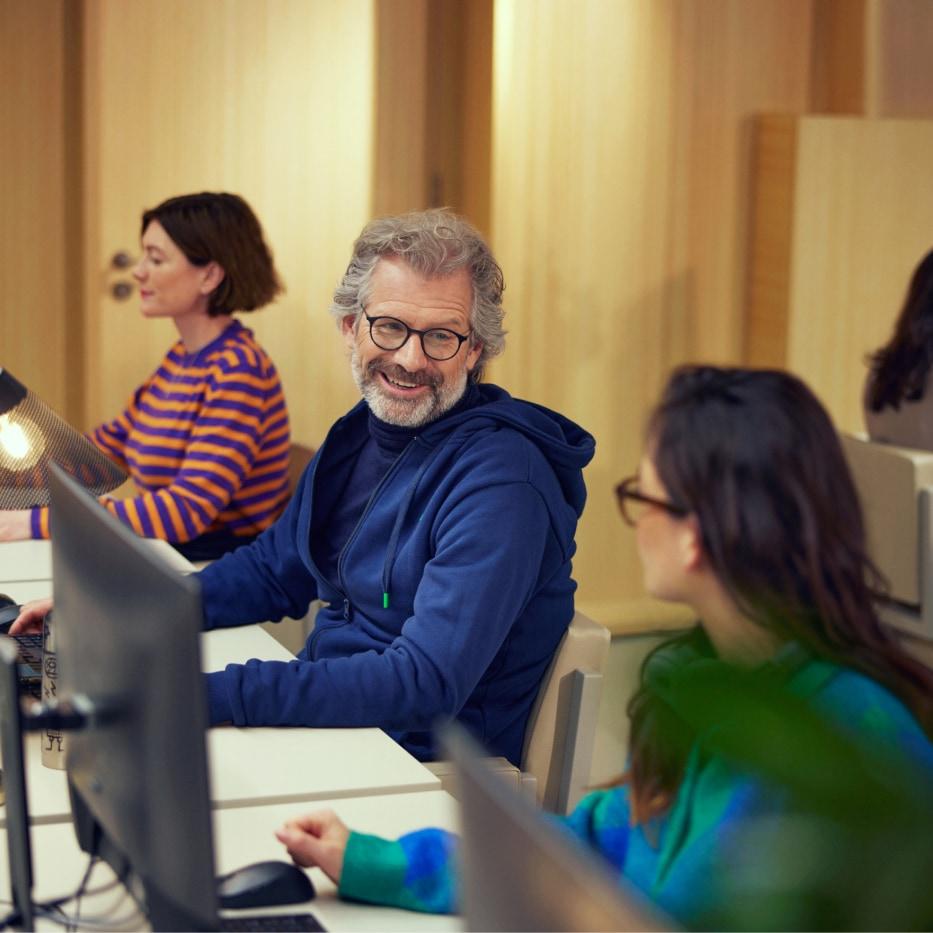 Smiling man with glasses working on computer in a modern office setting with colleagues.