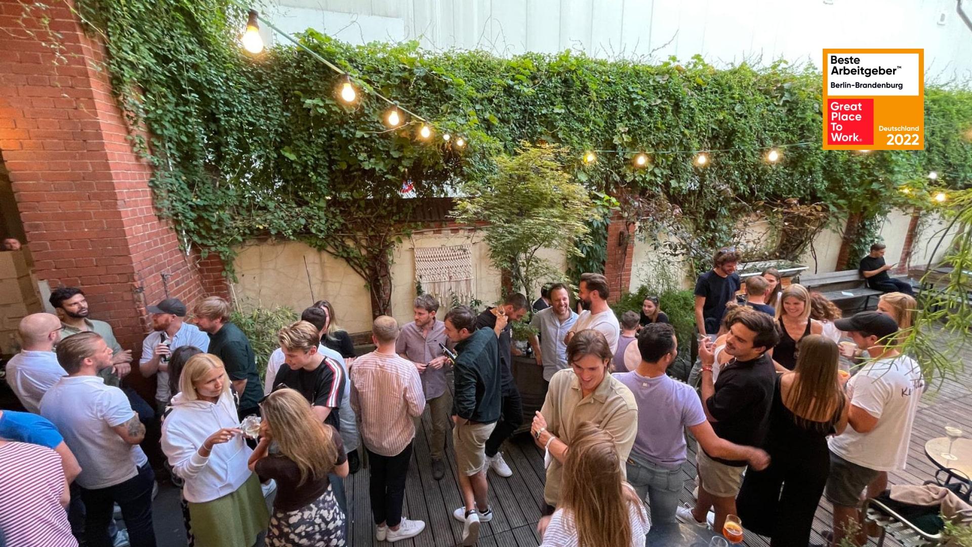 Gruppe von Menschen, die auf einer Außenterrasse mit Lichterketten und Grünanlagen sozialisiert.