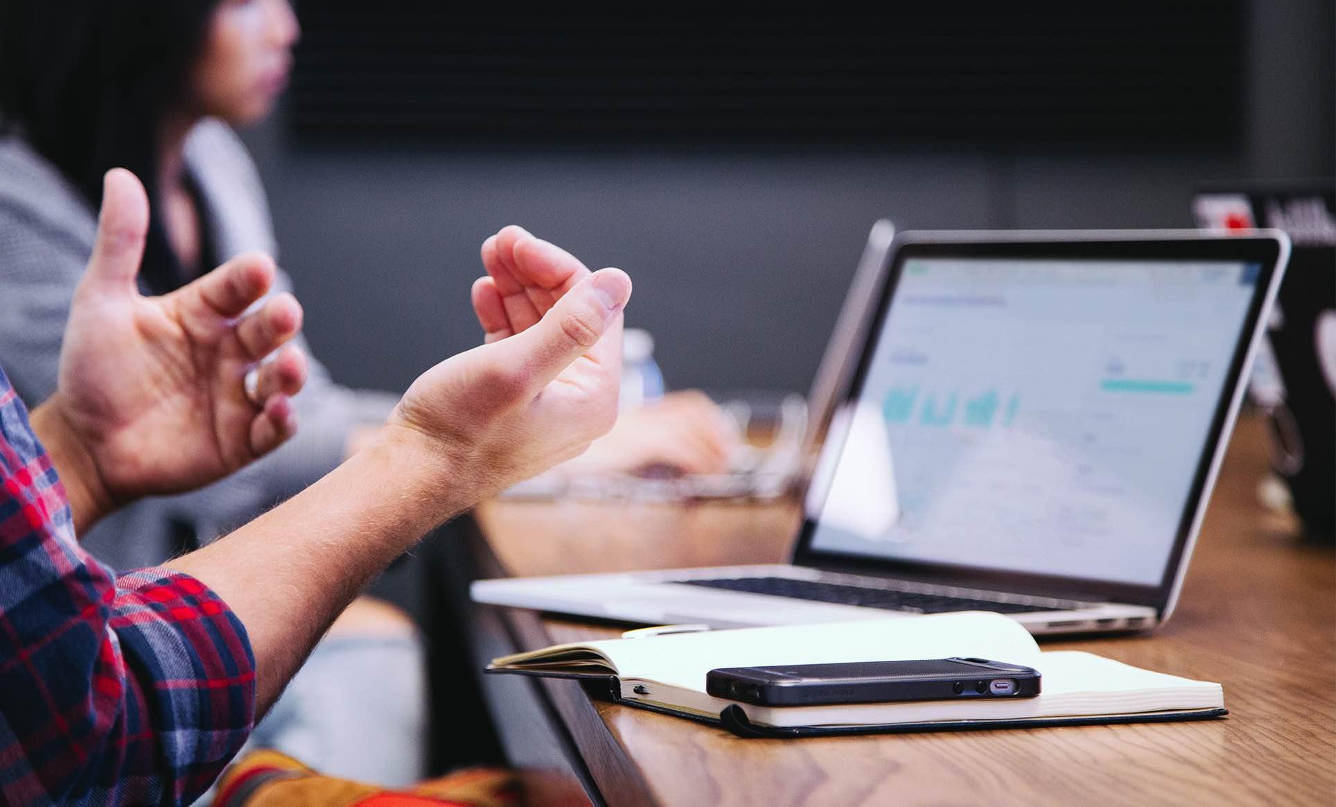 Personne gesticulant lors d'une réunion avec un ordinateur portable affichant des graphiques et un smartphone sur la table.
