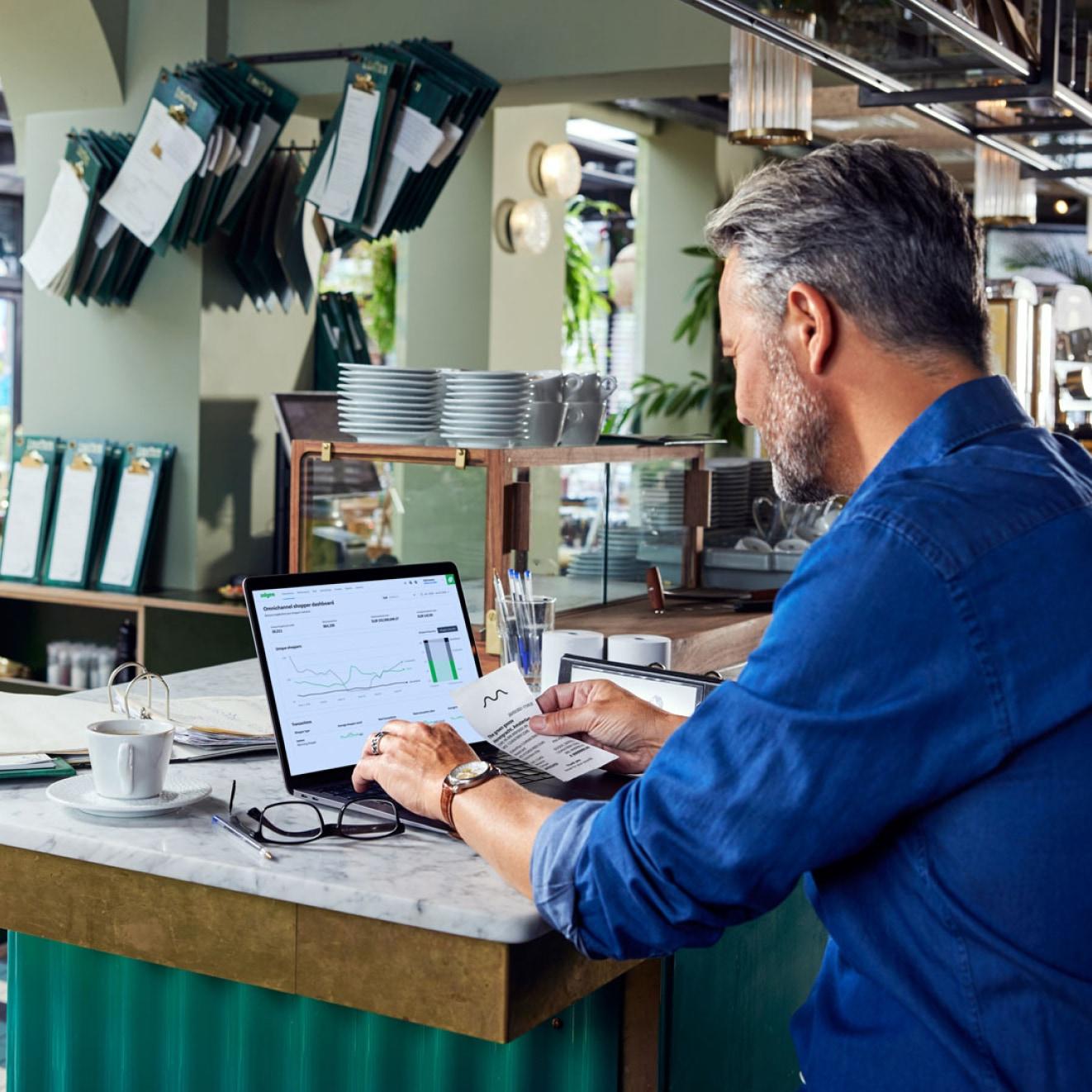 Man in cafe analyzing sales data on laptop screen with Adyen analytics visible.