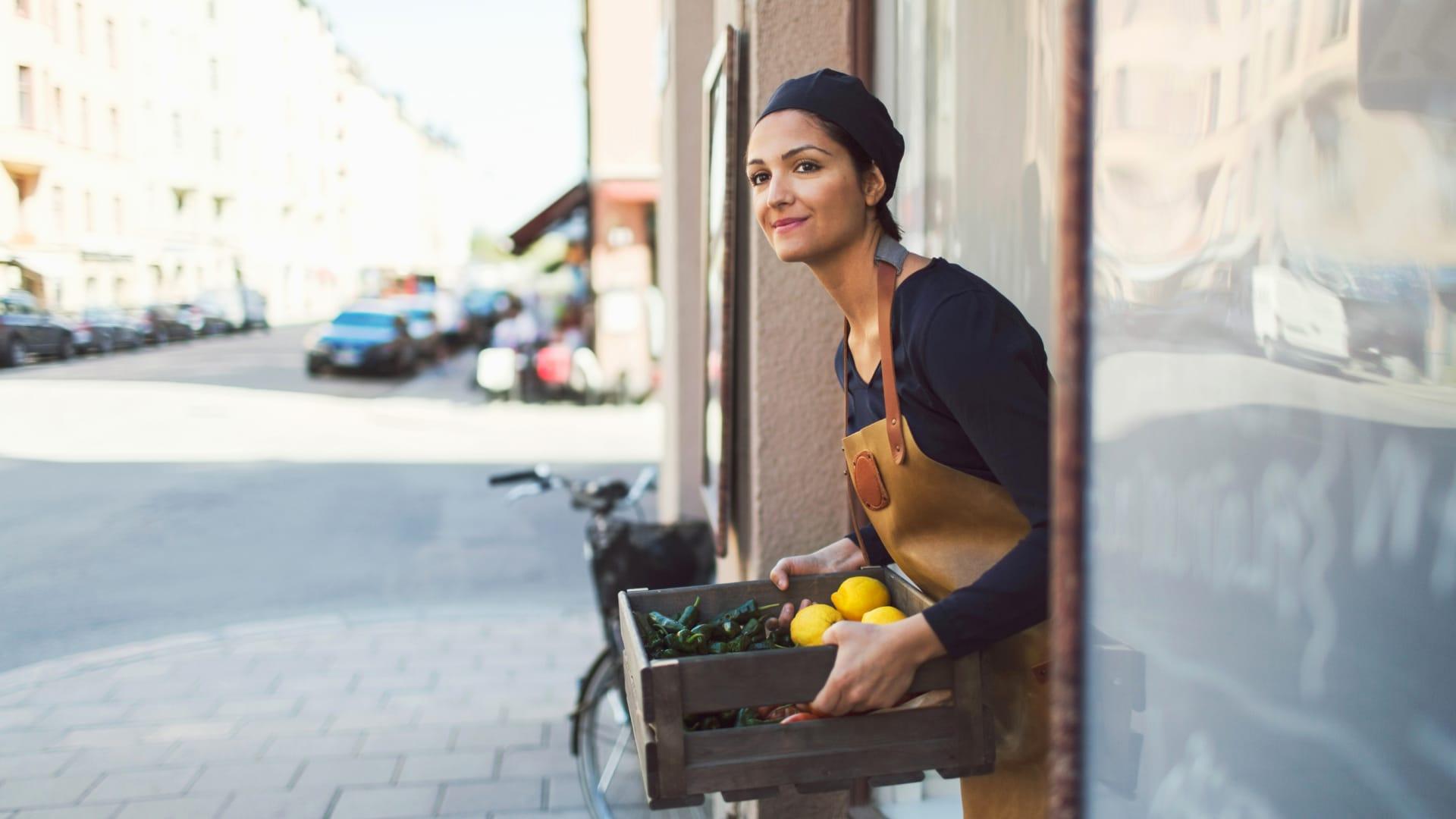 Woman in apron holding a crate of fresh lemons and vegetables outside a shop.