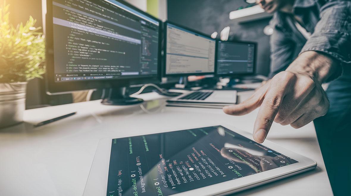 Persona interactuando con código en una tableta con varias pantallas de computadora en el fondo.