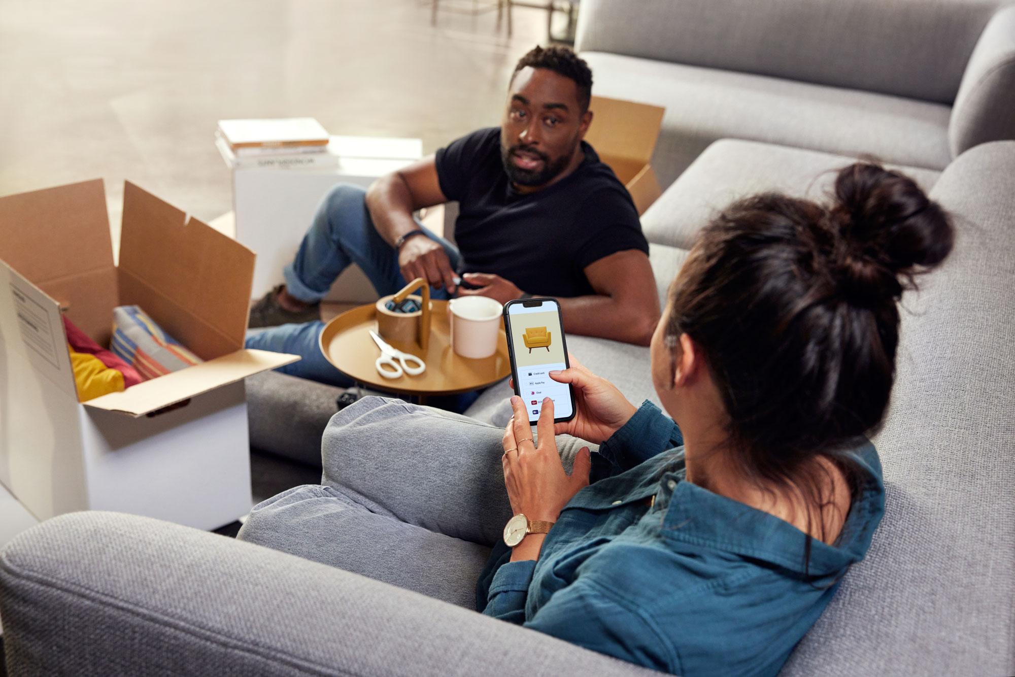 A couple sitting on a couch and shopping for furniture on their phone.