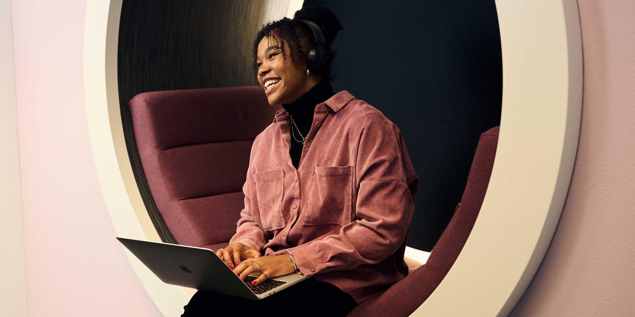 A person sitting and smiling while working on a laptop in a modern office environment.