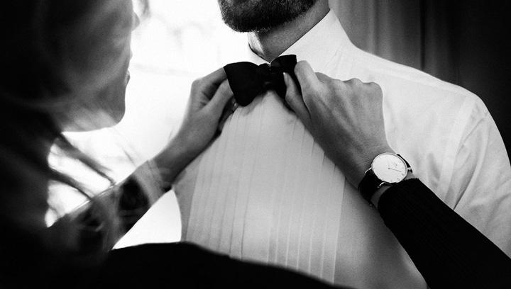 Black and white photo showing a person adjusting a bow tie on a man in a tuxedo with a watch.