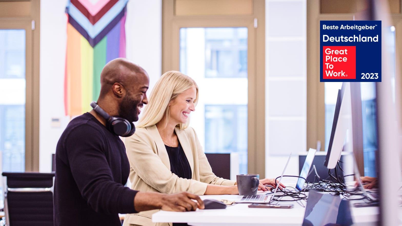Zwei Profis lächeln und arbeiten an Computern in einem Büroumfeld mit einem 'Great Place To Work 2023' Preis im Hintergrund.