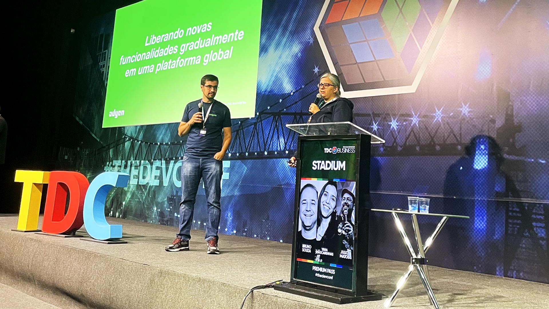 Dois palestrantes apresentam em uma conferência com slide Adyen ao fundo