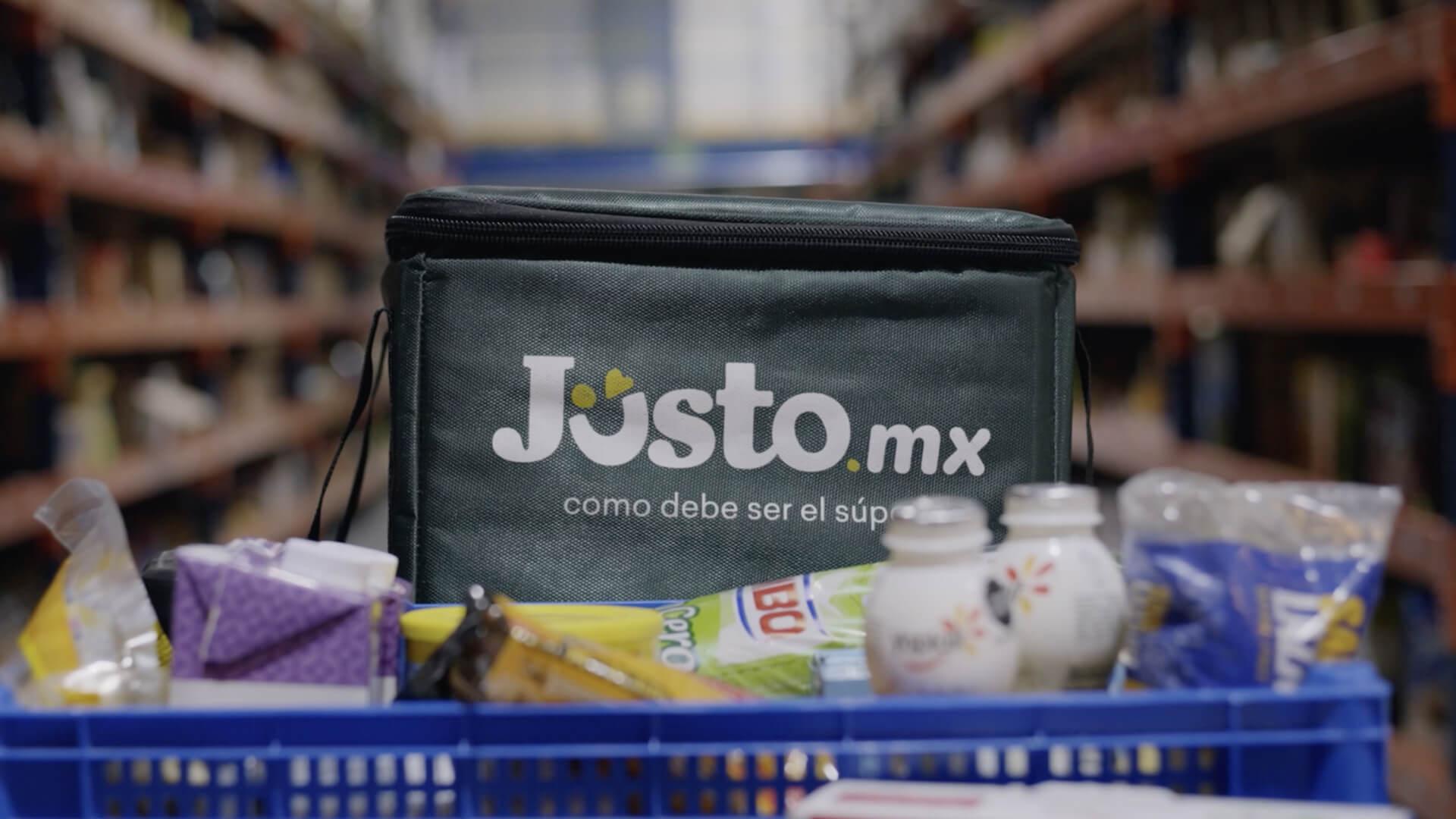 Artículos de alimentación en una cesta azul con una bolsa de reparto con la marca Justo.mx.