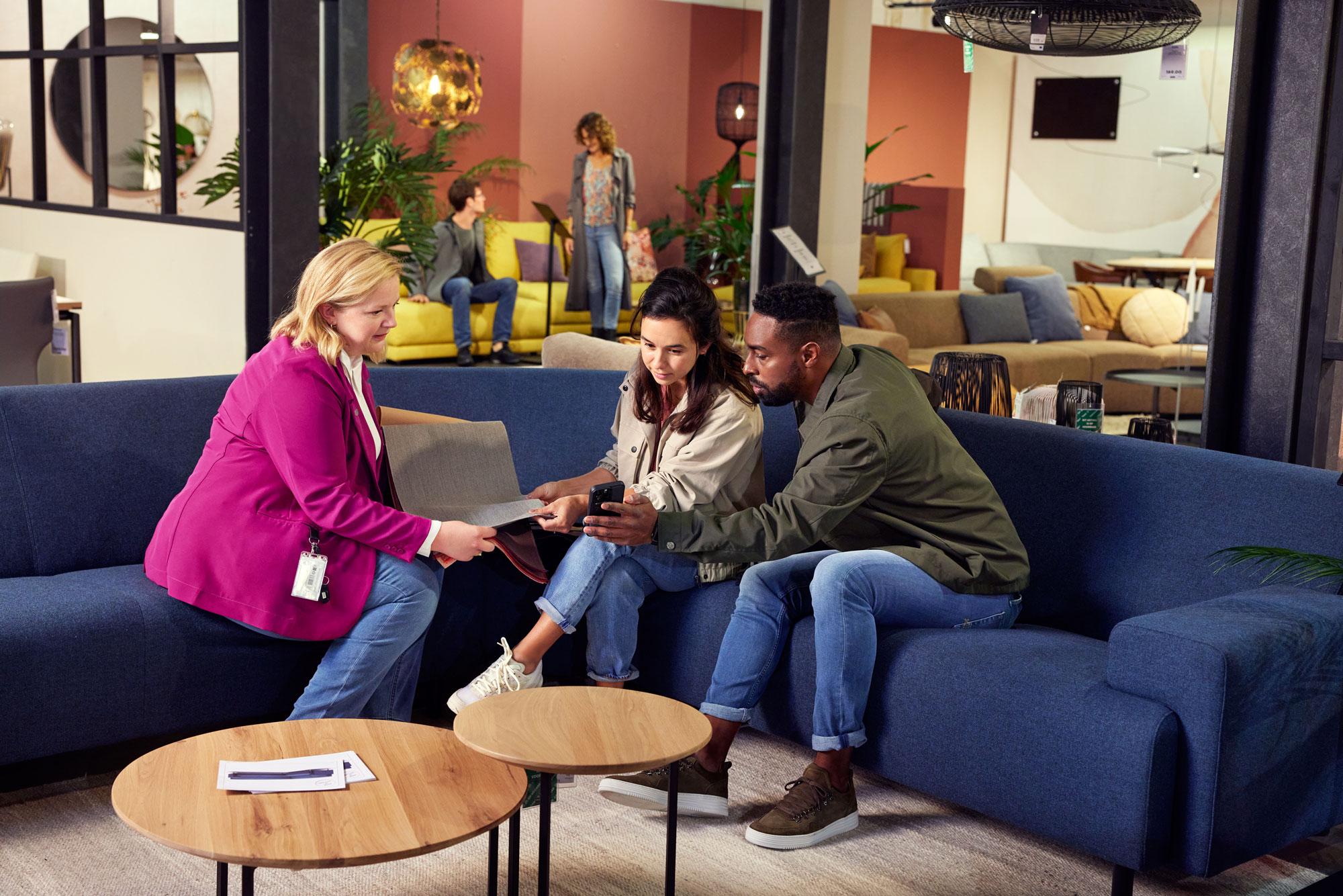 A couple in a furniture store sitting on a couch and discussing furniture products with an employee.