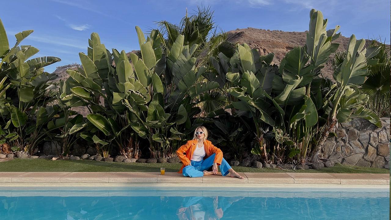 Woman in orange jacket and blue pants sitting by a pool with tropical plants in the background.