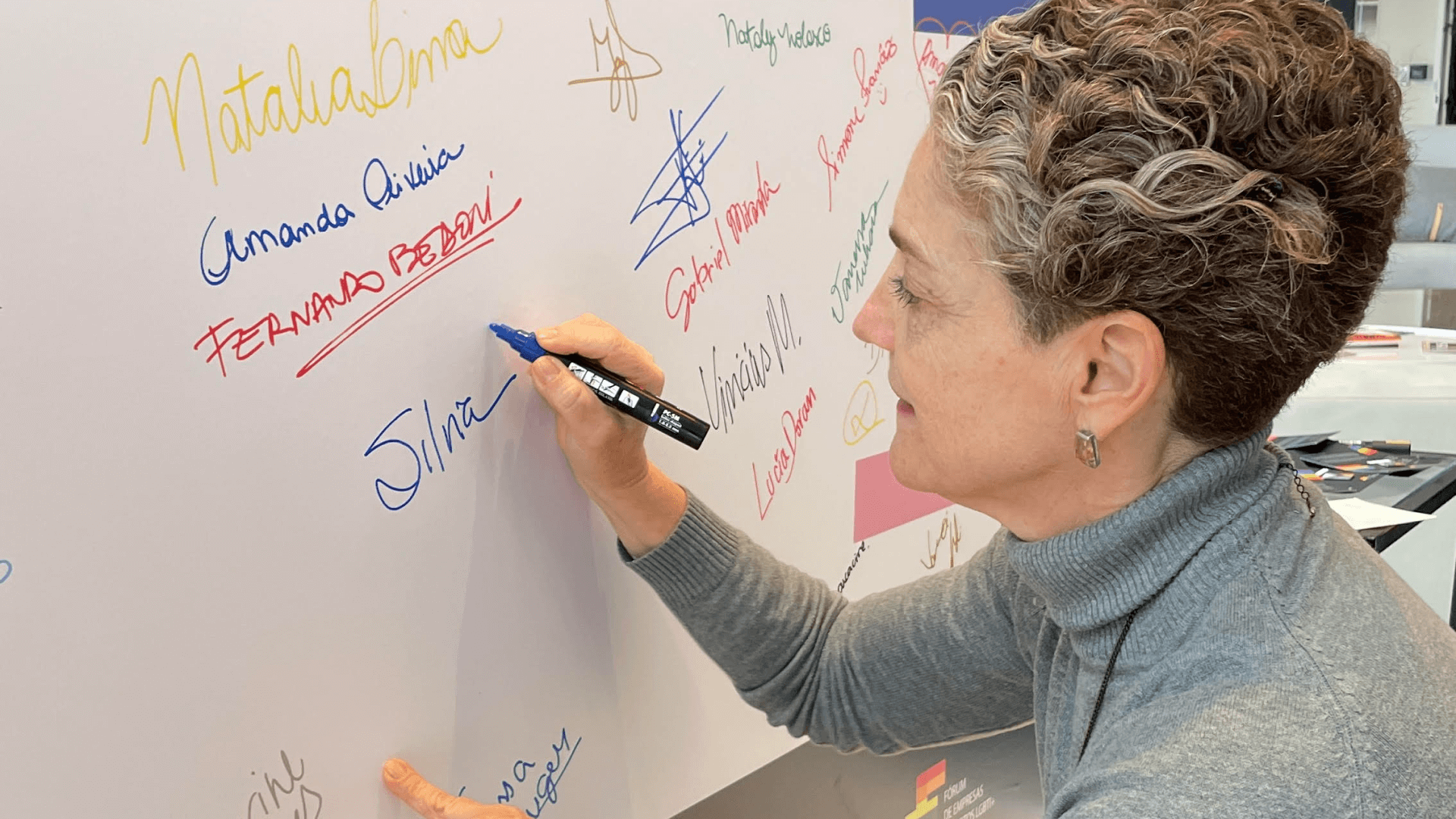 Pessoa assinando seu nome em um quadro branco ao lado de outras assinaturas.