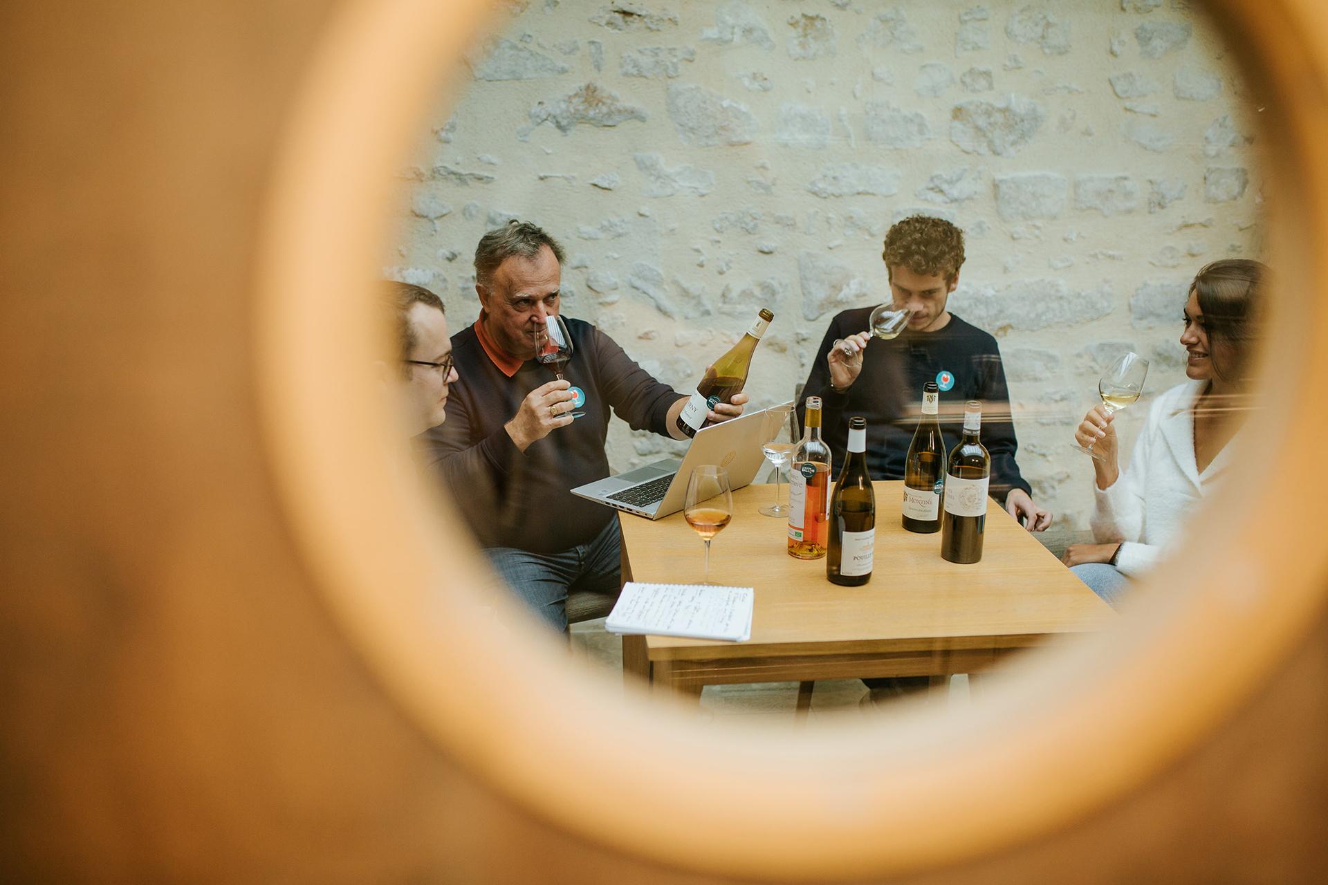 Groupe de personnes dégustant du vin autour d'une table avec un ordinateur portable et des bouteilles de vin dessus.