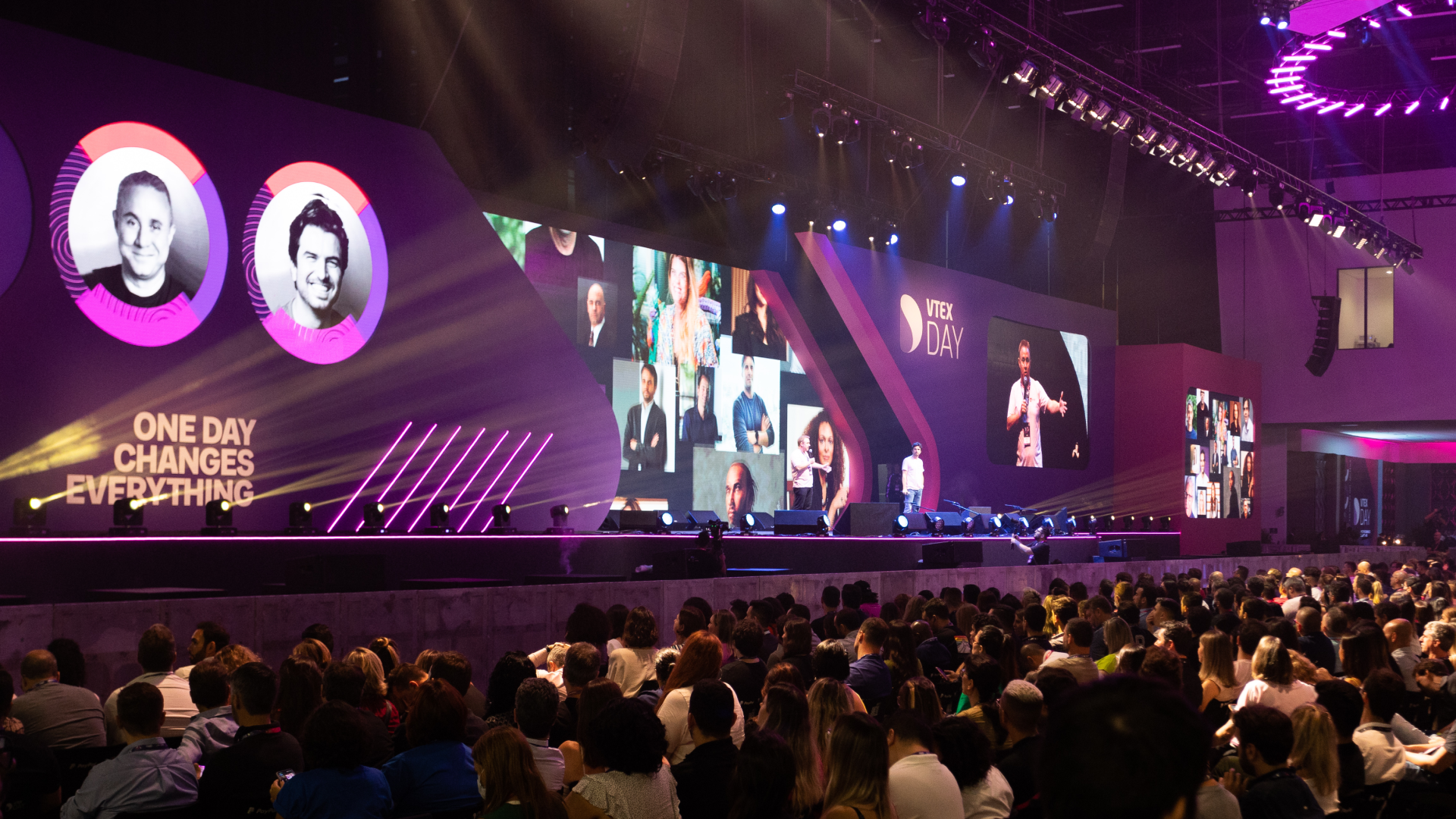 Público no evento VTEX DAY com palestrantes apresentados em telas grandes do palco
