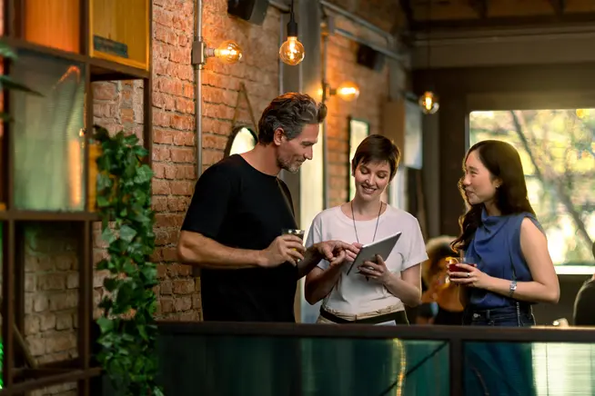 Dos mujeres y un hombre mirando una tableta en un restaurante y discutiendo suscripciones.