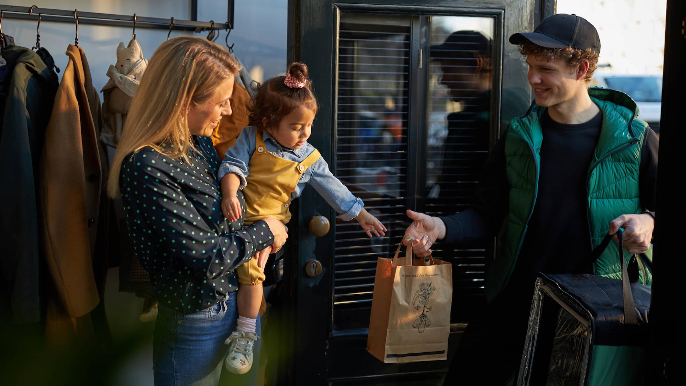 Woman and child receiving a shopping bag from a smiling delivery person at the doorway.