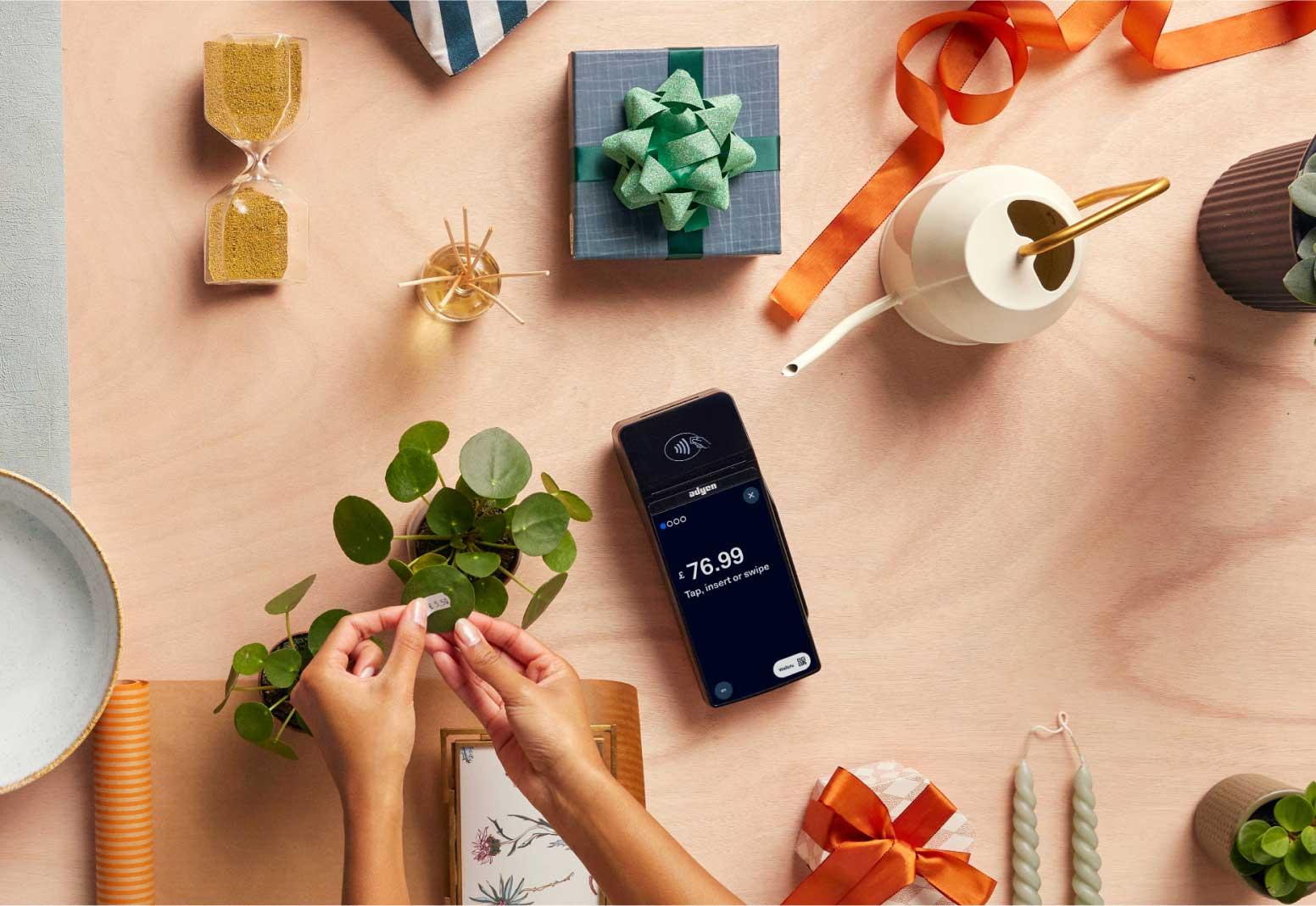 Hands wrapping a plant stem with a bow, next to a smartphone showing Adyen payment of $76.99.