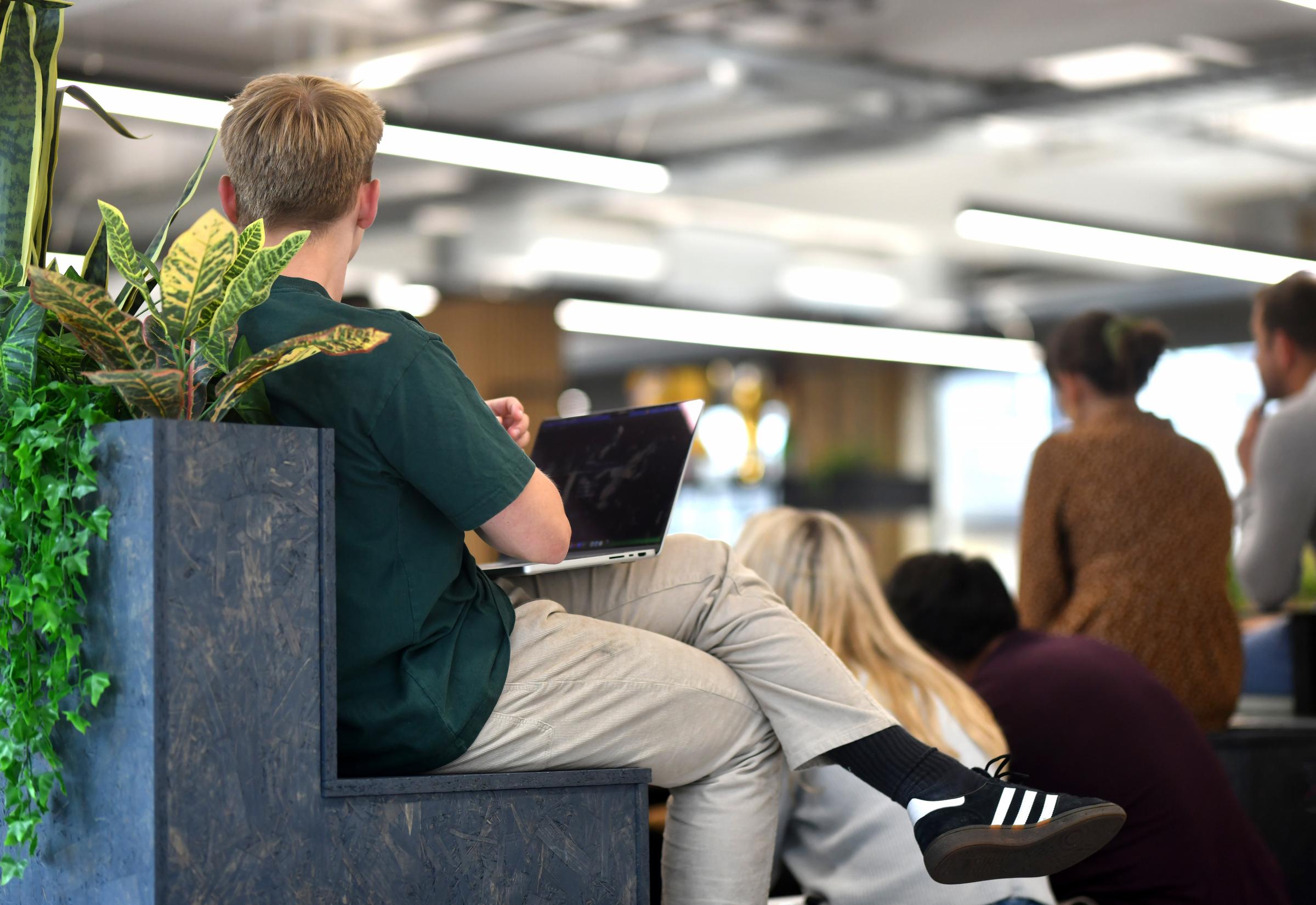 A man sitting on a bench behind this laptop in an office with people around him