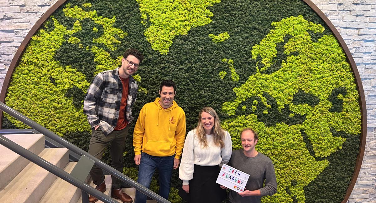 Four smiling individuals standing in front of a green wall with the shape of a globe.