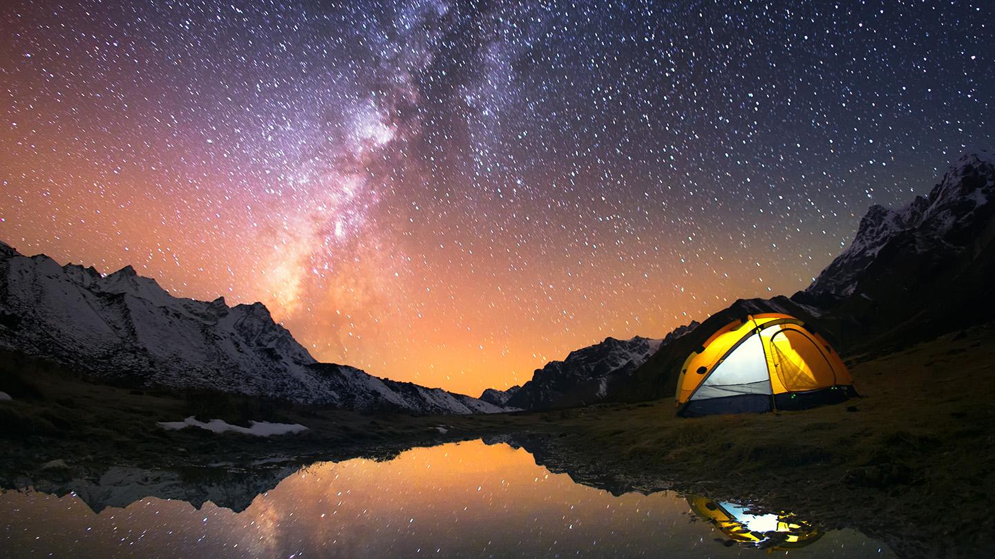 Tente éclairée sous un ciel étoilé avec la Voie lactée et des reflets de montagnes sur l'eau.