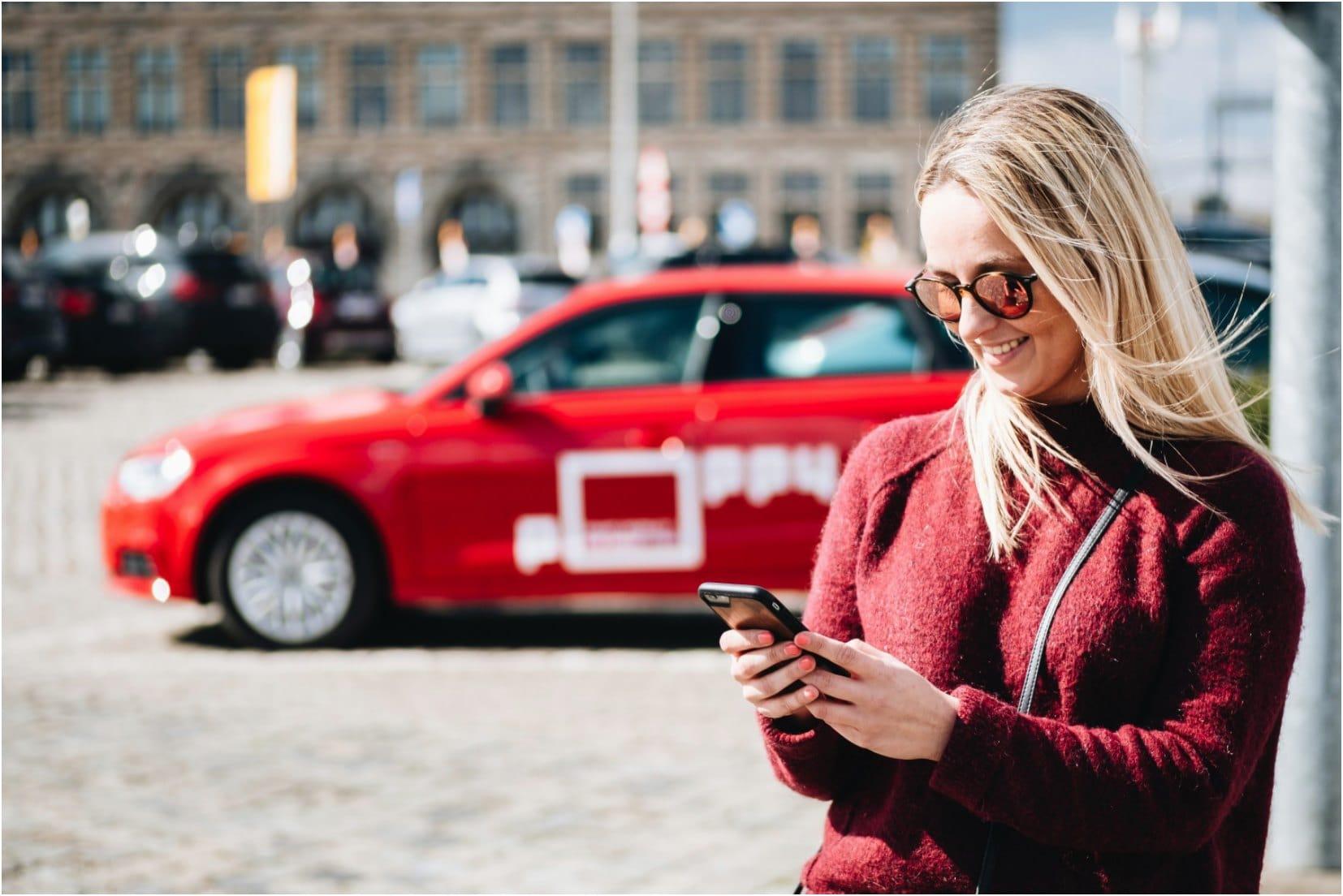 Femme souriante avec des lunettes de soleil utilisant son smartphone avec une voiture rouge en arrière-plan.